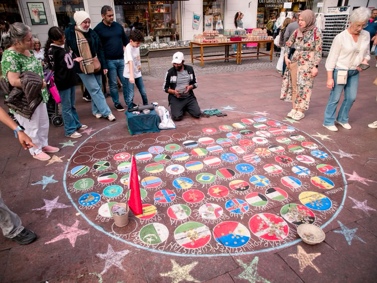 A group of people stand around a circular chalk art display on the ground, featuring various colorful designs and flags. A person is kneeling in the center of the circle, and there are stars drawn around the perimeter.