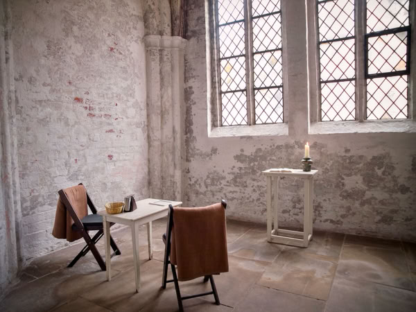 A small, rustic room with two wooden chairs and a table, each draped with a brown cloth. A candle is lit on a stand near a large window with a grid pattern.