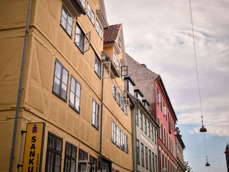 Colorful buildings with multiple windows line a street, with a cloudy sky and hanging streetlights above.