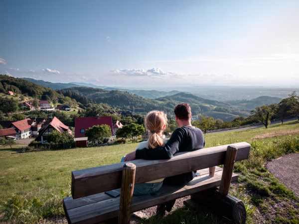 A couple sitting on a bench overlooking a scenic landscape with hills, houses, and a clear sky.