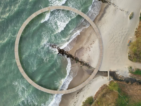 Aerial view of a circular pier over the ocean, with waves crashing against it, surrounded by sandy beach and greenery.
