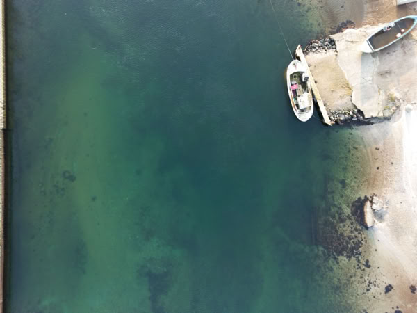 Aerial view of a small boat docked at a pier on a body of water, with a sandy shore and some rocks visible nearby.