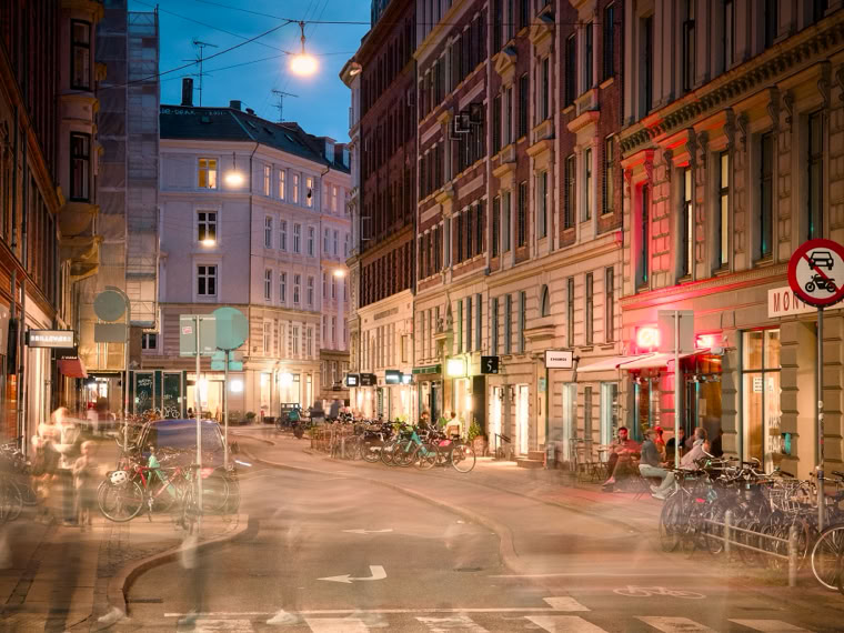 A city street scene at dusk with blurred motion of people and bicycles, surrounded by tall buildings with illuminated windows and streetlights.