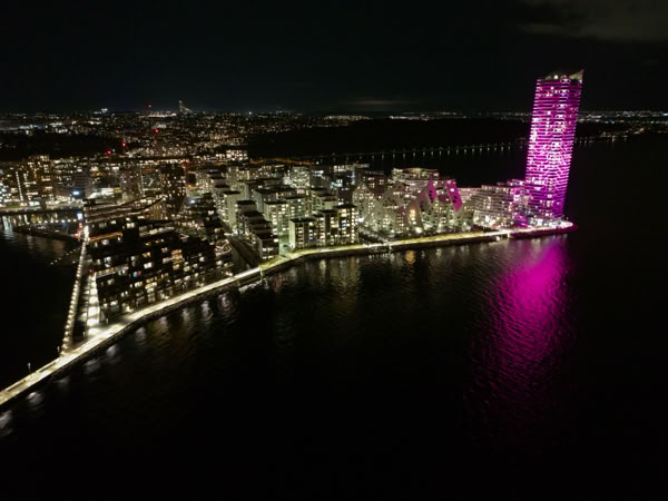 Aerial view of a cityscape at night with buildings illuminated, including a tall building lit in pink, reflecting on the water.