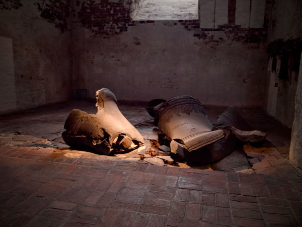 Broken, large bell pieces lying on a brick floor in a dimly lit room with rough, textured walls.