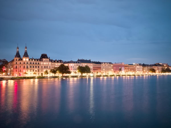 A waterfront cityscape at dusk with illuminated buildings reflecting on the water.