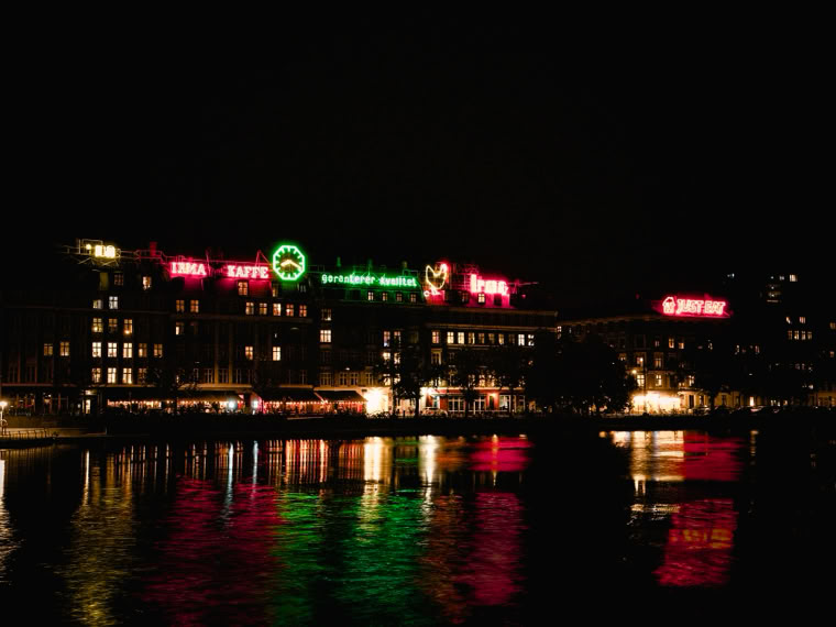 Cityscape at night with illuminated buildings and colorful neon signs reflecting on the water.