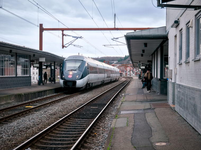 A train approaching a platform at a train station with overhead wires and a few people waiting.