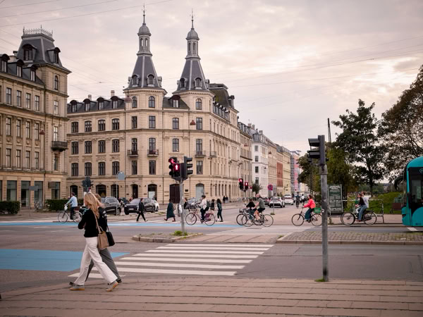 A city street scene with people walking and cycling, featuring historic buildings with ornate architecture and pointed towers in the background, and a tram on the right side.