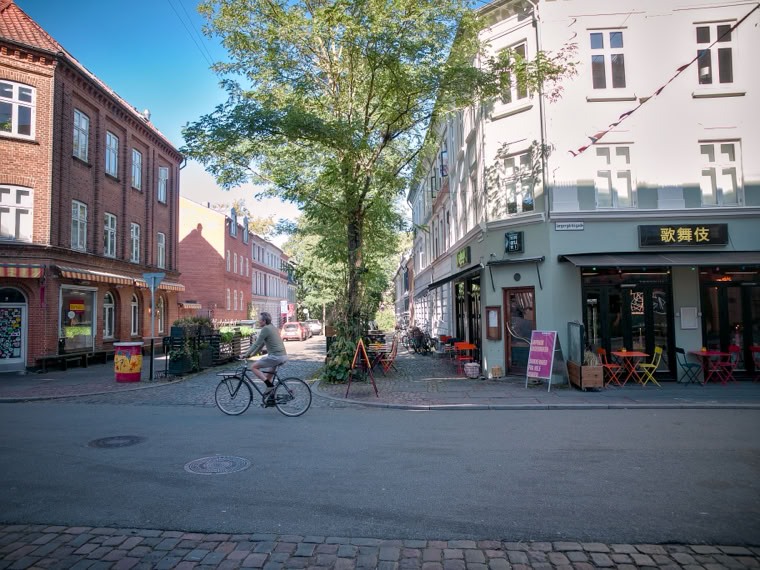 A street scene with a person riding a bicycle, buildings on either side, a tree in the center, and a café with outdoor seating on the right.