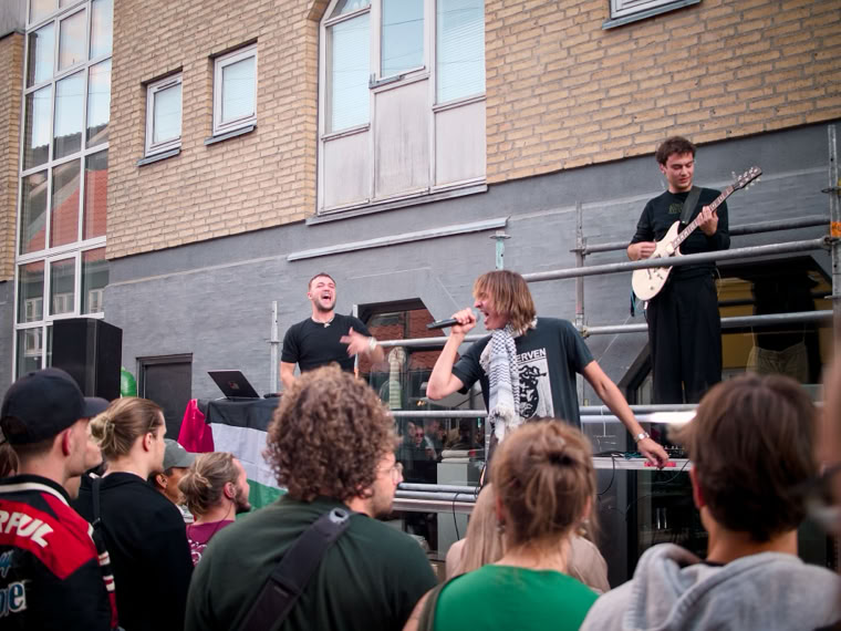 A group of people gathered outdoors in front of a building, with two men performing on a raised platform, one singing into a microphone and the other playing a guitar.