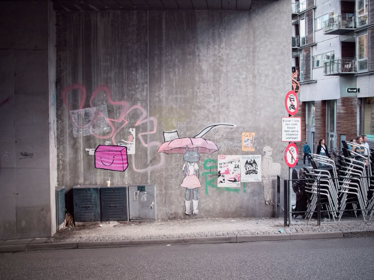 Street art on a concrete wall depicting a figure holding a pink umbrella, surrounded by graffiti and posters. Nearby are stacked chairs and traffic signs.