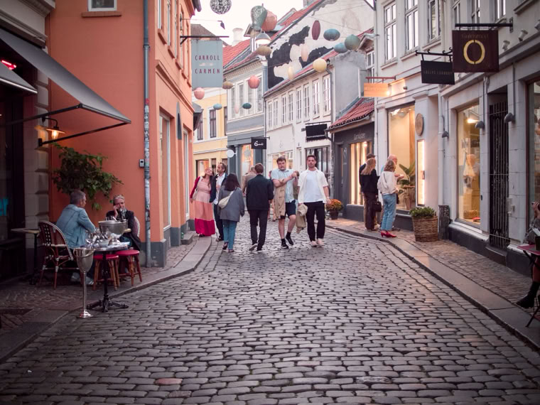 A cobblestone street lined with colorful buildings, outdoor seating at a café, and several people walking.