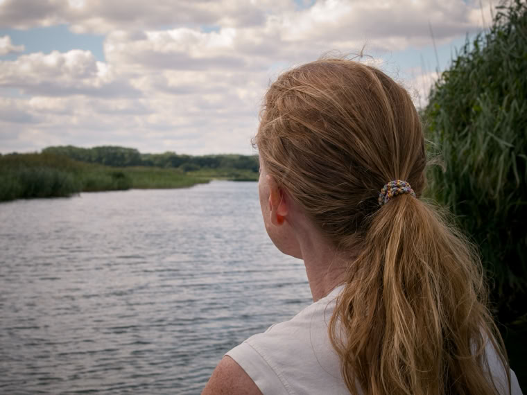 A person with long hair in a ponytail is looking out over a river, surrounded by greenery and under a partly cloudy sky.