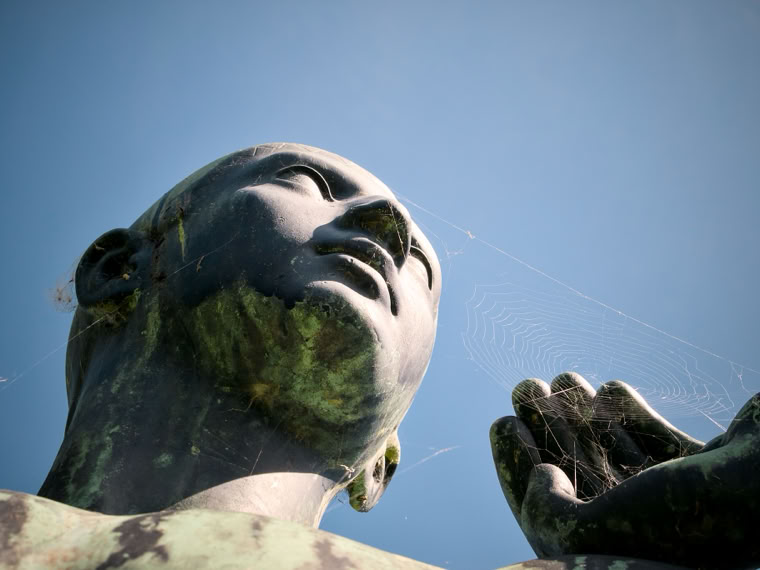 A low-angle view of a large statue with a serene expression, one hand raised, against a clear blue sky.