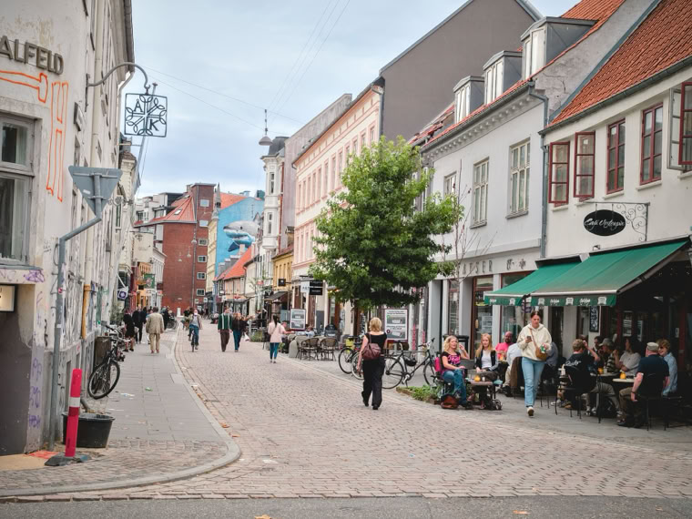 A cobblestone street lined with buildings, people walking, and a group sitting outside a cafe under a green awning. Bicycles are parked along the street, and a tree is in the center.