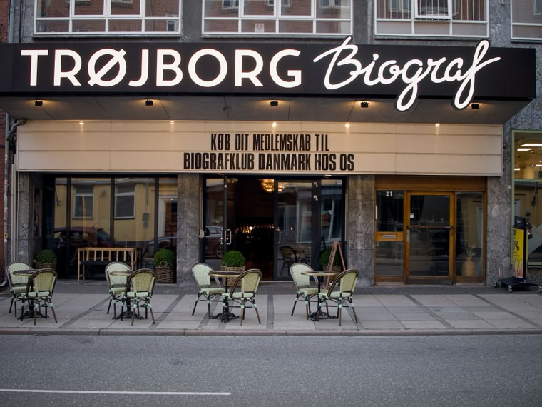 Exterior of a cinema with the sign "TRØJBORG Biograf" above the entrance, featuring a marquee and outdoor seating with tables and chairs on the sidewalk.