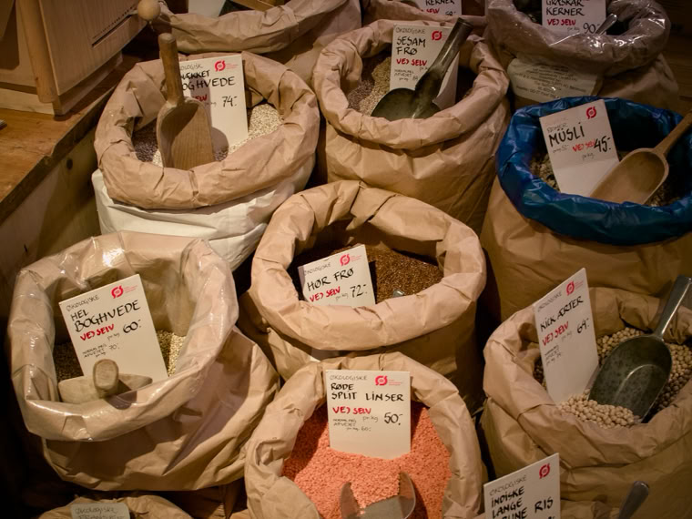 Bags of various grains and legumes with scoops and price tags displayed in a market setting.