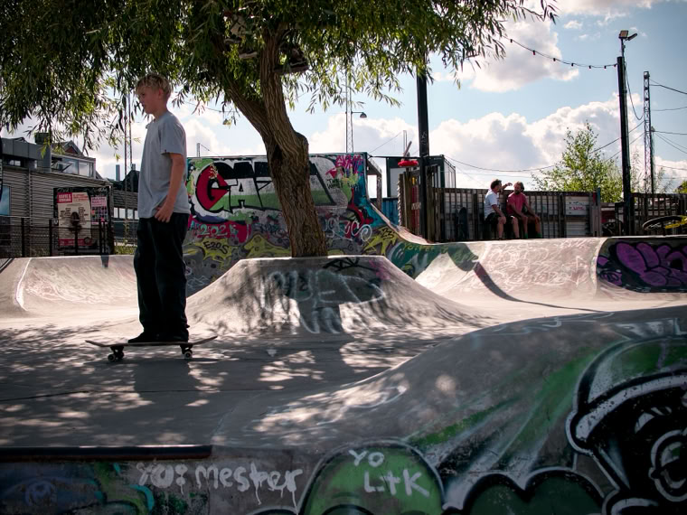 A person skateboarding in a graffiti-covered skate park with ramps and a tree in the background under a partly cloudy sky.