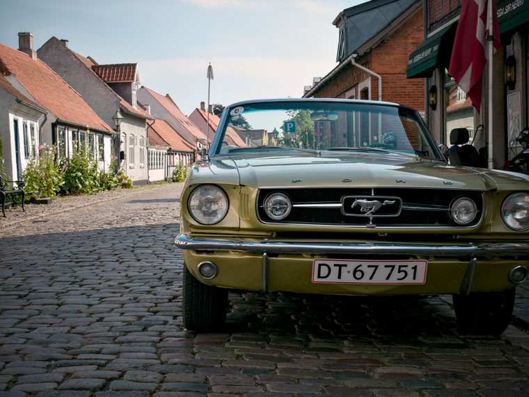 A vintage yellow car parked on a cobblestone street lined with quaint houses and shops.
